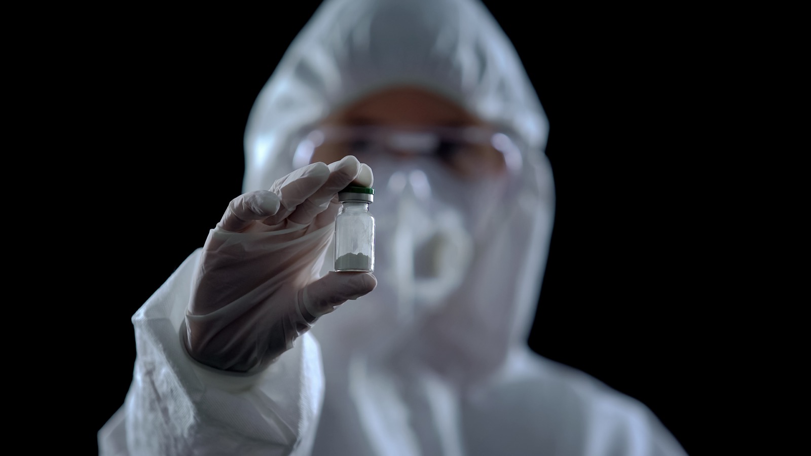 Person in full hazmat protective suit holding a single vial against a dark background