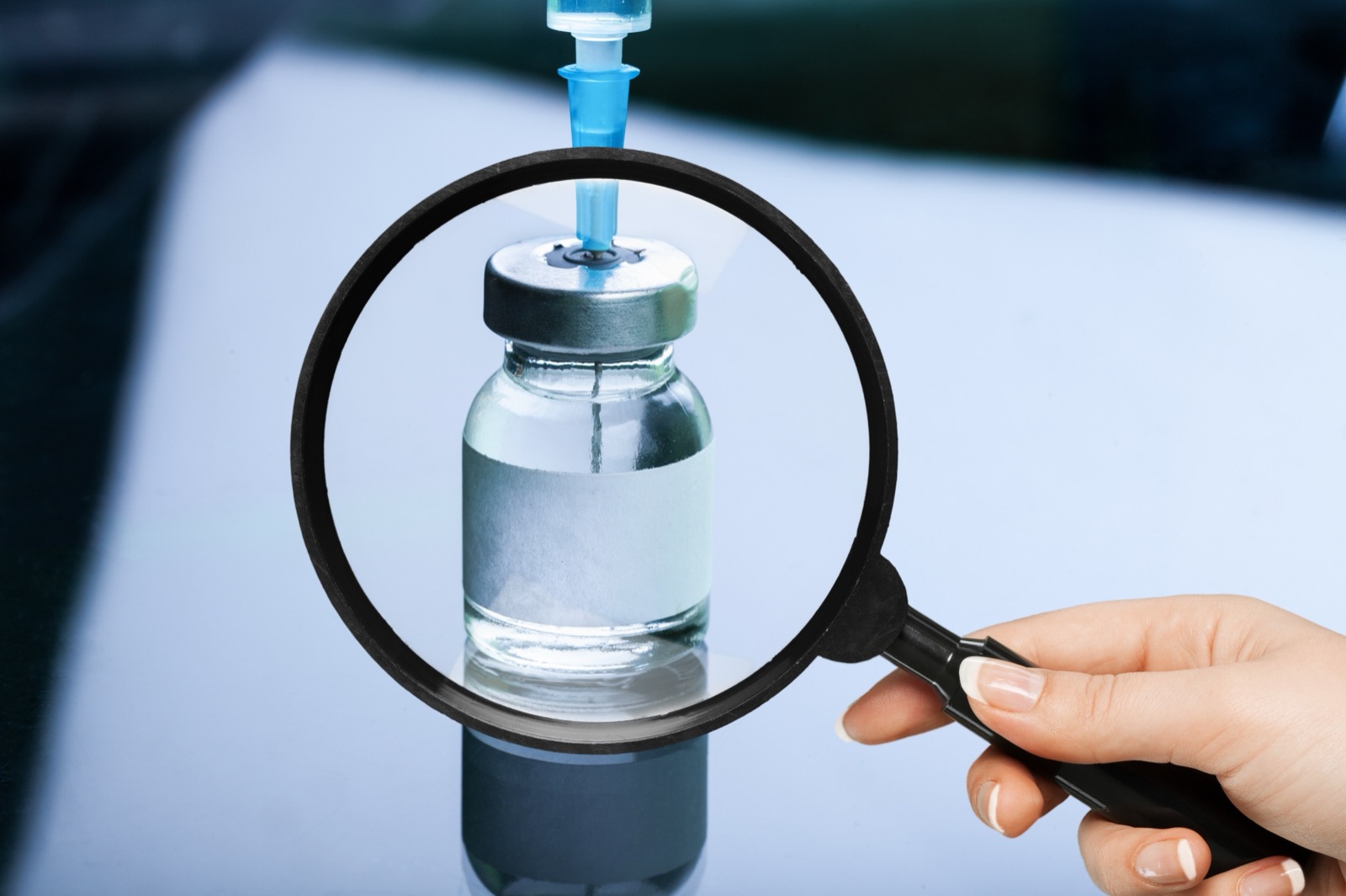 Magnifying glass held up to examine a small pharmaceutical vial, with a syringe resting beside it